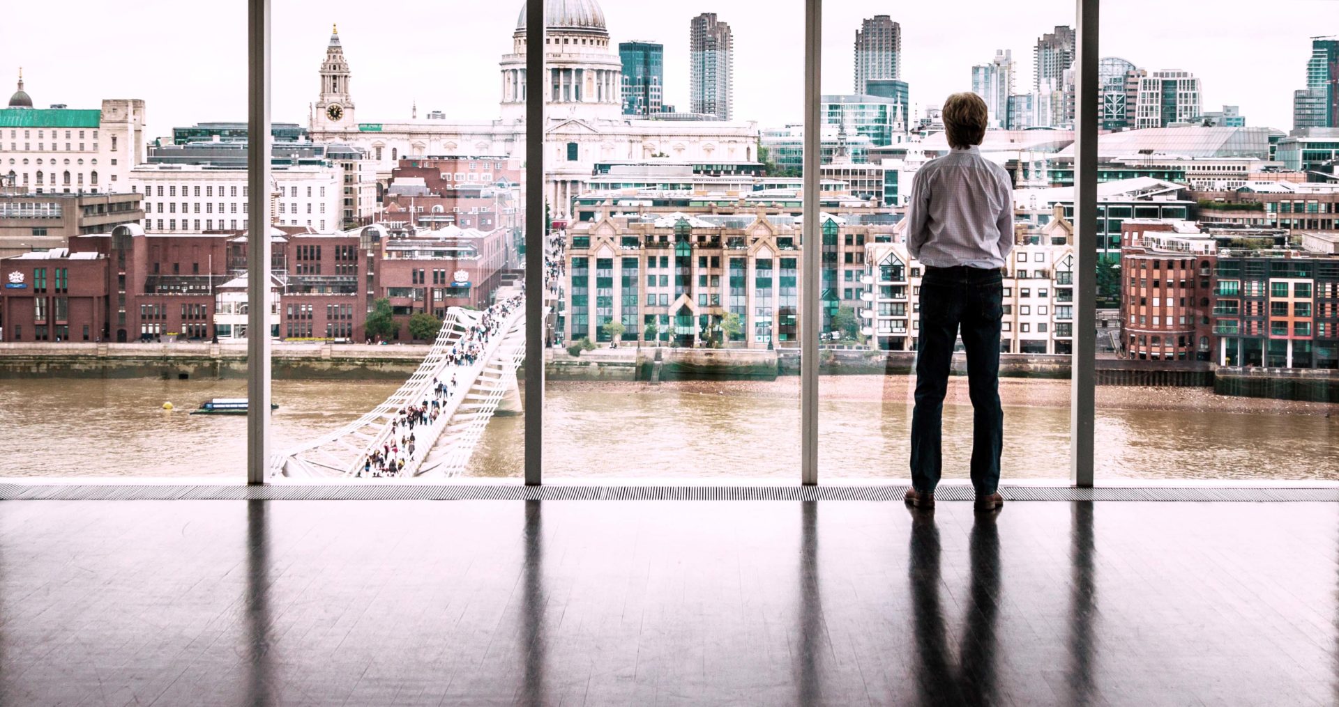Rear view image of a businessman looking out at the modern London skyline from high up in his office building. He is looking across the river Thames, across the Millennium Bridge to St Paul's cathedral and the high rise skyscrapers of the City of London. He has his hands folden in front of him. Horizontal colour image with copy space.